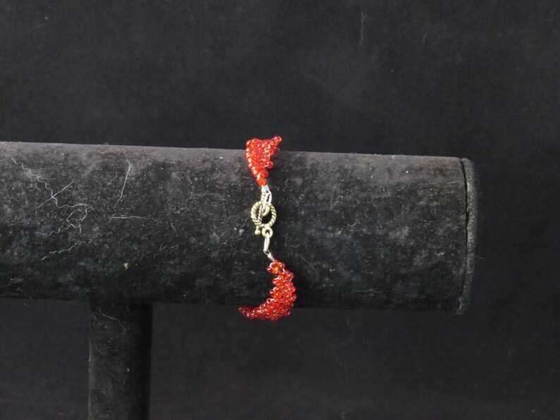 Puede incluir: Una pulsera de cuentas rojas con un cierre plateado. La pulsera est&aacute; hecha de peque&ntilde;as cuentas rojas redondas y se muestra sobre un soporte negro sobre un fondo oscuro. La pulsera mide aproximadamente 18 cm de largo.