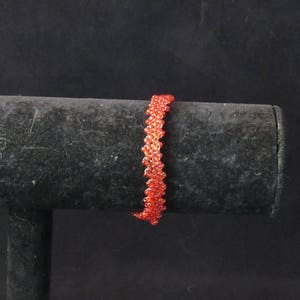 Puede incluir: Una pulsera de cuentas en tonos naranja y rojo, exhibida sobre un soporte negro. La pulsera est&aacute; hecha de peque&ntilde;as cuentas redondas, creando una apariencia texturizada y tejida. El fondo es negro liso.