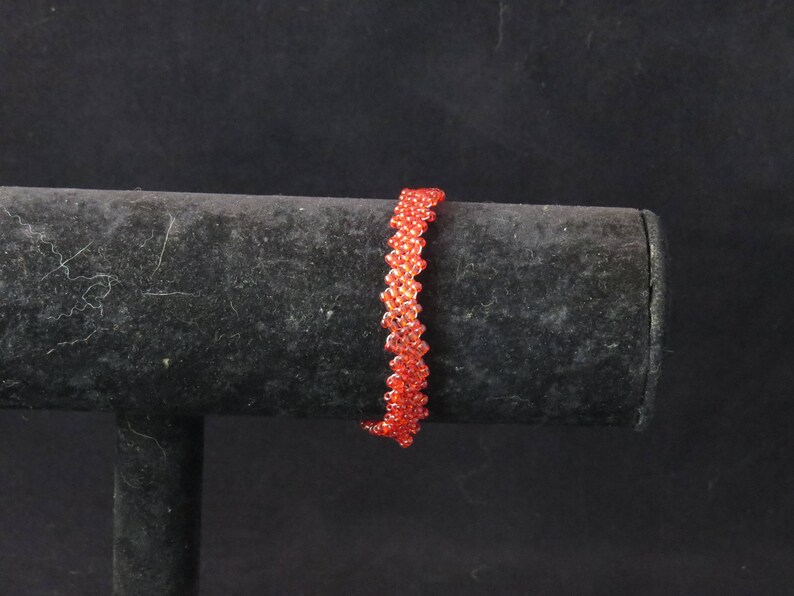 Puede incluir: Una pulsera de cuentas en tonos naranja y rojo, exhibida sobre un soporte negro. La pulsera est&aacute; hecha de peque&ntilde;as cuentas redondas, creando una apariencia texturizada y tejida. El fondo es negro liso.