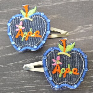 Può includere: Due fermagli per capelli a forma di mela in denim blu con dettagli ricamati, tra cui la parola "Mela" e piccoli fiori. I fermagli sono su uno sfondo in legno.