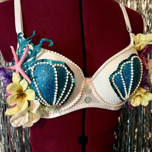 Puede incluir: Sujetador blanco con copas de concha de mar de brillo azul y turquesa, decorado con cuentas de perlas, flores rosas y amarillas y una estrella de mar.