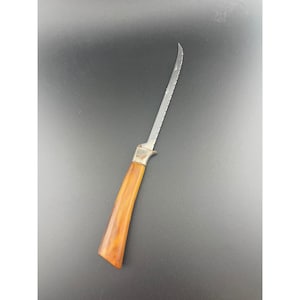May include: A vintage carving knife with a curved, serrated stainless steel blade. The handle is a warm, amber-toned material, possibly wood or a composite, with a silver-coloured bolster. The knife is designed for slicing meats.