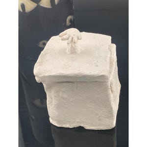 May include: A square, off-white ceramic box with a textured, matte finish. The lid features a small, sculpted hand-shaped handle at the center. The box has irregular, hand-molded edges and a solid, opaque appearance, presenting a tactile, clay-like surface.
