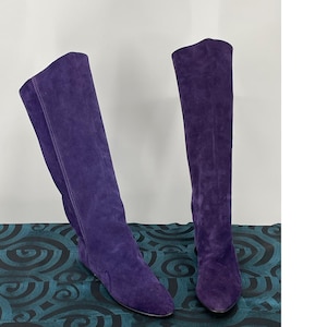 May include: A pair of knee-high boots crafted from deep purple suede. These boots feature a simple, pull-on design with a rounded toe, a flat sole, and vertical seam detailing running along the outer side of the shaft for a clean, uniform look.
