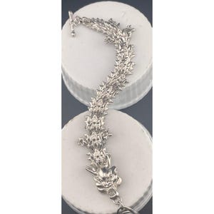 Può includere: Un braccialetto argentato con un design a forma di drago. Il braccialetto ha una chiusura a testa di drago dettagliata e una struttura a catena con una trama a spillo. La chiusura a levetta è visibile in alto. Il braccialetto è esposto su una superficie bianca.