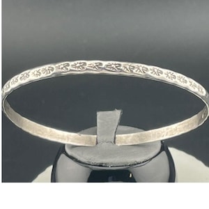 Può includere: Un braccialetto rigido argentato con un motivo floreale sul bordo superiore. Il braccialetto è esposto su un supporto nero su sfondo scuro. Il motivo floreale si ripete su tutto il braccialetto.