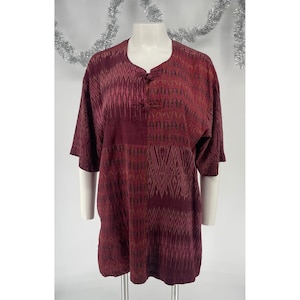Puede incluir: Una túnica de manga corta en tonos burdeos profundo y rojo oscuro, con un complejo patrón geométrico estilo ikat. La prenda presenta un cuello redondo con dos cierres de palanca cubiertos de tela en la parte central delantera.