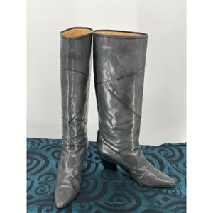 May include: A pair of knee-high boots in a smooth, dark grey finish. The boots feature a pointed toe, a low block heel, and a series of diagonal stitched seams across the shaft that create a geometric pattern. The interior lining is tan.