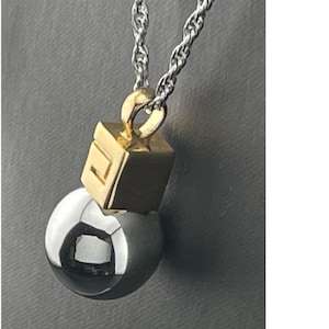 May include: A silver chain necklace with a pendant featuring a gold-colored square and a silver sphere. The gold square has a geometric design. The sphere is reflective, with a dark gray color.