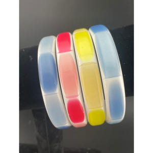 May include: Four colorful, rectangular bangles. Each bangle has a white base with a gradient of color on top. The colors are blue, pink, yellow, and blue. The bangles are displayed on a black surface.