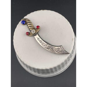 May include: A silver-toned dagger-shaped brooch with ornate floral engravings on the blade. The hilt features a blue gemstone at the top, red gemstones on either side, and a clear gemstone in the center. The brooch is set on a white, round surface.