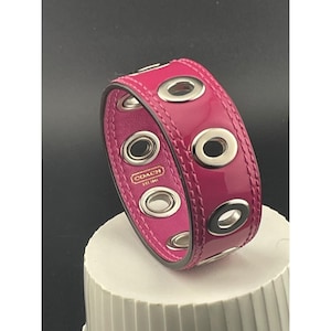 May include: A vibrant, hot pink Coach bracelet featuring silver eyelets. The bracelet has a leather interior and is displayed on a white surface. The Coach logo is visible on the inside of the bracelet.