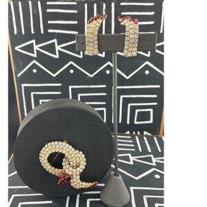 Könnte beinhalten: Ein goldfarbenes Schmuckset bestehend aus einem Paar Creolen und einer runden Brosche. Jedes Teil ist mit klaren weißen Strasssteinen besetzt und mit kleinen, tiefroten tropfenförmigen Steinen in einer strukturierten Metallfassung verziert.