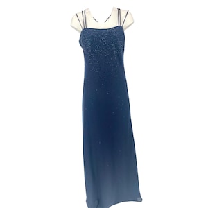 May include: A navy blue formal gown with a beaded bodice and spaghetti straps. The floor-length dress features a subtle ombre effect, transitioning from a darker shade at the top to a lighter hue at the hem. The dress is displayed on a mannequin.