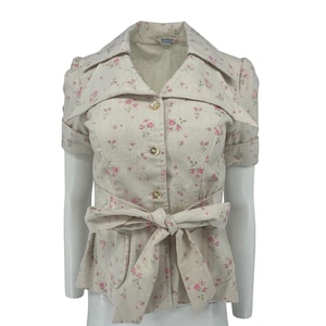 May include: A beige short-sleeved jacket with a floral pattern of pink roses. The jacket features a notched collar, gold buttons, and a matching fabric belt tied in a bow at the waist. The jacket is displayed on a mannequin.