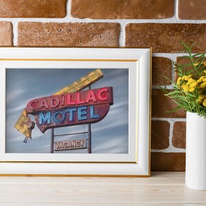 K&ouml;nnte beinhalten: Ein gerahmtes Foto eines alten Cadillac Motel Neonschilds mit roter und blauer Schrift vor bew&ouml;lktem Himmel, pr&auml;sentiert in einem wei&szlig;en Rahmen mit Goldrand, platziert neben einer wei&szlig;en Vase mit gelben Blumen.