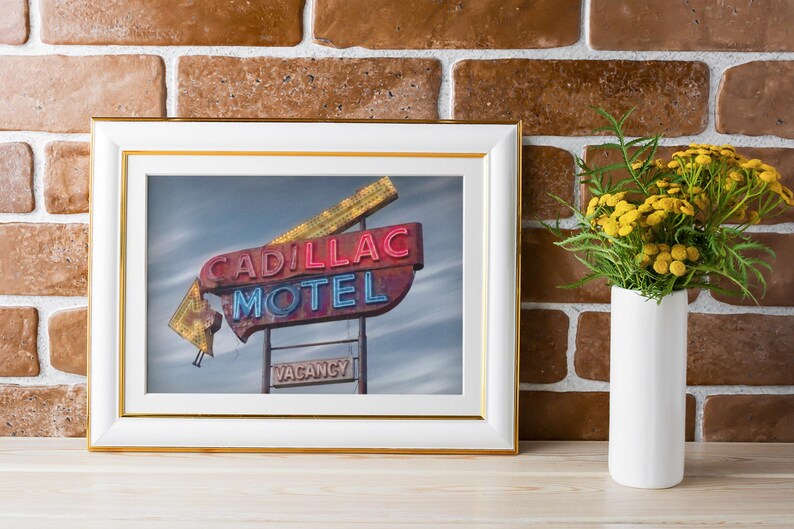 K&ouml;nnte beinhalten: Ein gerahmtes Foto eines alten Cadillac Motel Neonschilds mit roter und blauer Schrift vor bew&ouml;lktem Himmel, pr&auml;sentiert in einem wei&szlig;en Rahmen mit Goldrand, platziert neben einer wei&szlig;en Vase mit gelben Blumen.