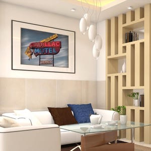 K&ouml;nnte beinhalten: Ein wei&szlig;es Sofa mit braunen und blauen Kissen steht vor einem Couchtisch aus Glas. Dar&uuml;ber h&auml;ngt ein gerahmter Druck mit einem Cadillac Motel Neonschild. Rechts befindet sich ein Raumteiler aus hellem Holz mit offenen Regalen f&uuml;r B&uuml;cher.