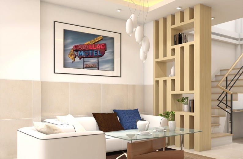 K&ouml;nnte beinhalten: Ein wei&szlig;es Sofa mit braunen und blauen Kissen steht vor einem Couchtisch aus Glas. Dar&uuml;ber h&auml;ngt ein gerahmter Druck mit einem Cadillac Motel Neonschild. Rechts befindet sich ein Raumteiler aus hellem Holz mit offenen Regalen f&uuml;r B&uuml;cher.