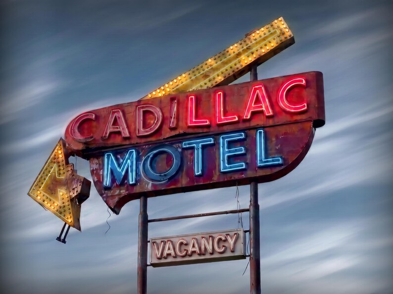 K&ouml;nnte beinhalten: Ein altes Cadillac Motel-Schild aus Metall mit roter Neon-Schrift f&uuml;r Cadillac und blauer Neon-Schrift f&uuml;r Motel. Das Schild enth&auml;lt einen gelben Pfeil, der nach unten zeigt, und ein kleineres rechteckiges Schild darunter mit der Aufschrift Vacancy.
