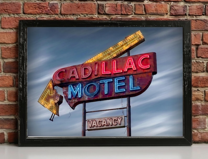 K&ouml;nnte beinhalten: Ein gerahmter Kunstdruck mit einem alten Cadillac Motel Neonschild. Das Schild zeigt die W&ouml;rter CADILLAC in rotem Neon, MOTEL in blauem Neon und ein kleineres VACANCY-Schild darunter. Ein gro&szlig;er gelber Neonpfeil zeigt vor einem bew&ouml;lkten Himmel nach unten.