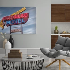 K&ouml;nnte beinhalten: Ein rechteckiger Wandkunstdruck mit einem verwitterten Neonreklameschild im Vintage-Stil, das die Aufschrift CADILLAC MOTEL in roten und blauen Buchstaben zeigt, sowie ein kleineres VACANCY-Schild darunter vor einem bew&ouml;lkten Himmel.