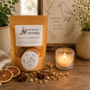 Könnte beinhalten: Ein brauner Papierbeutel mit der Aufschrift Sunday morning. Botanical Simmer Blend und einem Sichtfenster mit getrockneten Orangenscheiben. Das Etikett zeigt eine Vogelillustration und Text. Daneben liegen Orangenscheiben, Sternanis und eine brennende Kerze.