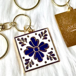 Puede incluir: Llavero dorado con un dije de diseño de azulejos azul y blanco. El dije presenta una flor estilizada con un contorno dorado. El llavero también incluye una etiqueta dorada con el texto "AKBALY ILLUSTRATION & DESIGN".