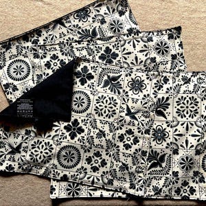 Puede incluir: Tres manteles individuales de tela con estampado en blanco y negro con respaldo de tela negro. Los manteles individuales presentan un diseño geométrico repetitivo con motivos florales. Los manteles individuales están apilados uno encima del otro y tienen una etiqueta que dice "Remarkable".