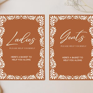 Puede incluir: Dos letreros marrones con bordes florales blancos y texto blanco. Los letreros dicen "Ladies Please Help Yourself Here's a Basket to Help You Along" y "Gents Please Help Yourself Here's a Basket to Help You Along".