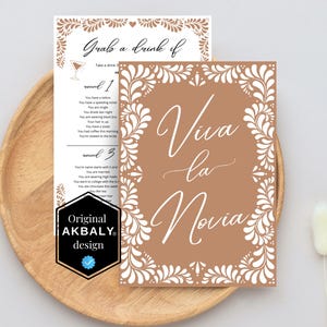 May include: A brown card with white floral designs around the edges. The card says "Viva la Novia" in white script. The card is on a wooden surface.
