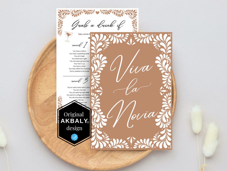 May include: A brown card with white floral designs around the edges. The card says "Viva la Novia" in white script. The card is on a wooden surface.