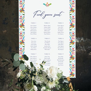 May include: A white seating chart with a colorful floral border. The chart lists guest names and table numbers for a wedding reception. The text "Find your seat" is written in blue cursive at the top of the chart.