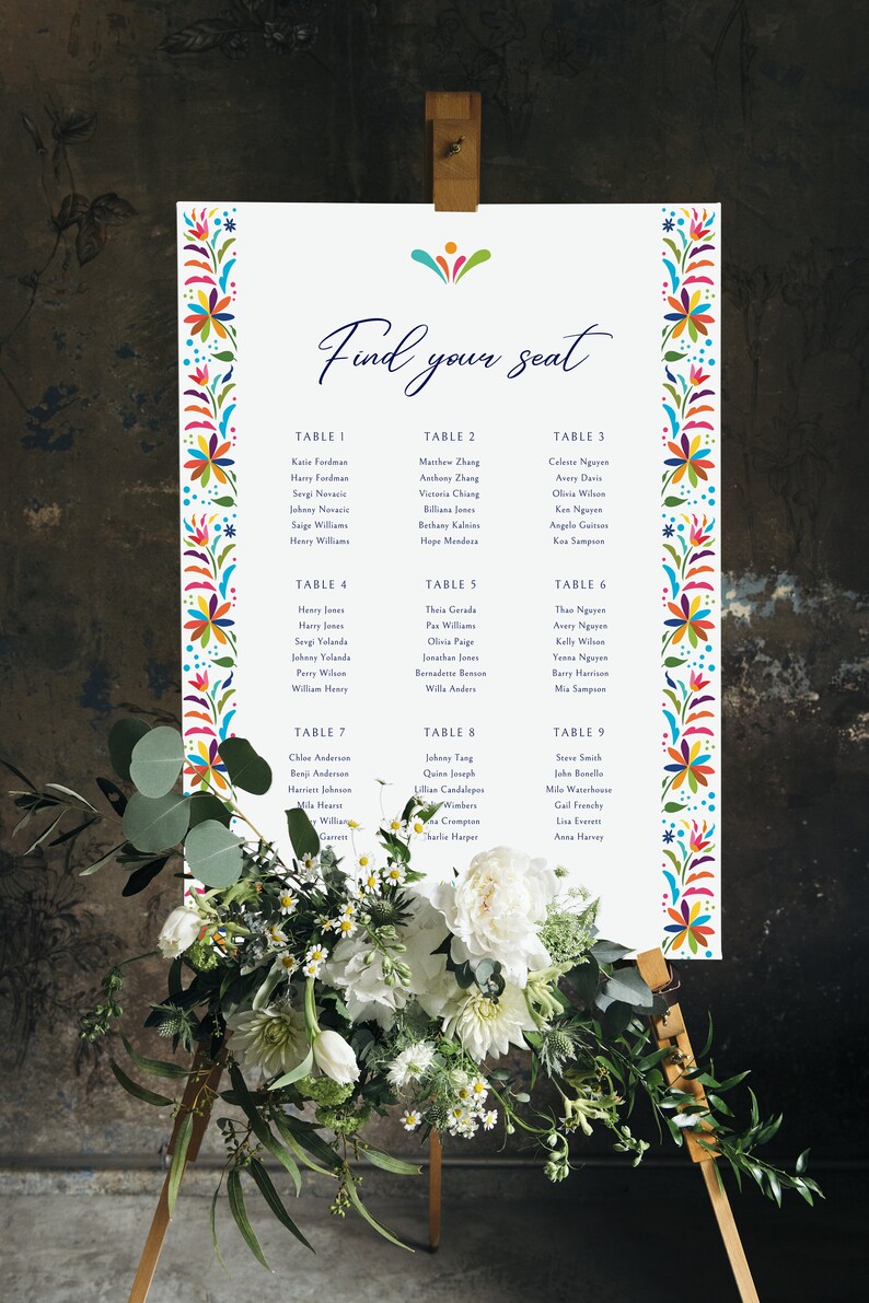 May include: A white seating chart with a colorful floral border. The chart lists guest names and table numbers for a wedding reception. The text "Find your seat" is written in blue cursive at the top of the chart.