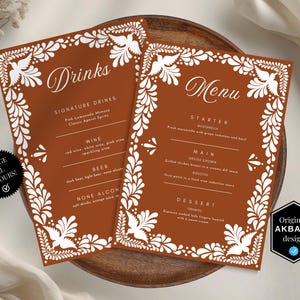 Puede incluir: Dos tarjetas de menú de color terracota con diseños florales y de pájaros blancos. Las tarjetas, etiquetadas como "Bebidas" y "Menú", presentan una lista de bebidas y una comida de tres platos. Las tarjetas están sobre un plato de madera.