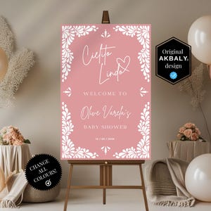 May include: A pink baby shower sign on a wooden easel, featuring white floral accents and the text "Cielto Linda" and "Welcome to Olive Varela's Baby Shower". The sign is surrounded by balloons and floral arrangements.