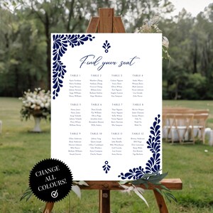 Peut inclure: Un plan de table de mariage blanc avec un motif floral bleu marine. Le plan est intitul&eacute; "Find your seat" et liste les noms des invit&eacute;s par num&eacute;ro de table. Le plan est affich&eacute; sur un chevalet dans un jardin.