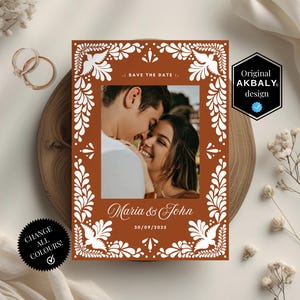 Może przedstawiać: Karta Save the Date ze zdjęciem pary, obramowana białymi kwiatowymi i gołębimi wzorami na brązowym tle. Karta zawiera imiona "Maria & John" i datę "30/09/2025". Projekt oznaczony jako "Original AKBALY design".