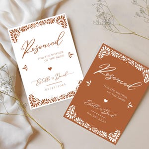 Puede incluir: Dos tarjetas de lugar de mesa de boda con un dise&ntilde;o floral blanco y el texto "Reservado para la madre de la novia" con los nombres de la pareja y la fecha de la boda. Las tarjetas est&aacute;n sobre un fondo marr&oacute;n claro.