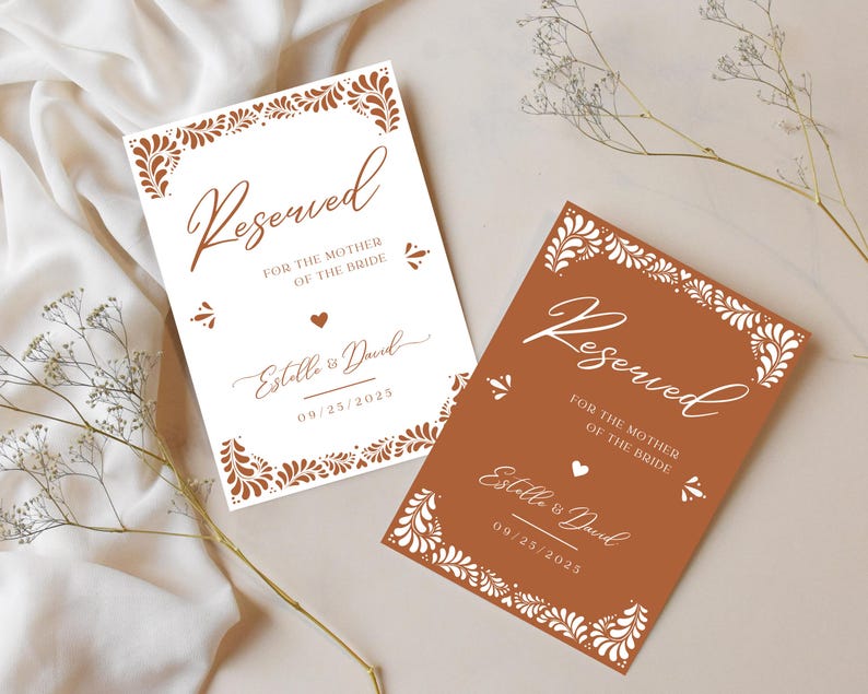 Puede incluir: Dos tarjetas de lugar de mesa de boda con un dise&ntilde;o floral blanco y el texto "Reservado para la madre de la novia" con los nombres de la pareja y la fecha de la boda. Las tarjetas est&aacute;n sobre un fondo marr&oacute;n claro.