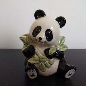 May include: A ceramic panda bear figurine with black and white coloring, holding green bamboo leaves. The panda has black ears, eyes, and paws, and is sitting down. The figurine is a decorative item.
