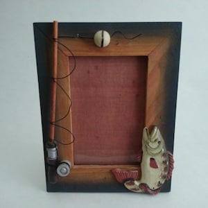 May include: Wooden picture frame with a fishing theme. The frame has a dark blue border and a light brown inner frame. A decorative fish is on the lower right corner. A fishing rod, bobber, and reel are attached to the frame.