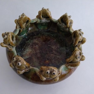 May include: A ceramic bowl with a circular design, featuring a brown and green glaze. The rim is adorned with several small, tan frog figurines. The bowl's interior has a dark, textured appearance, creating a unique decorative piece.