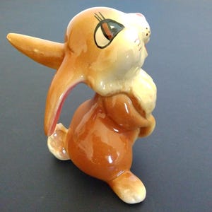 May include: A ceramic figurine of a rabbit in a standing pose. The rabbit is primarily brown with white and cream accents. It has large ears, a fluffy chest, and is painted with details like eyelashes and a pink inner ear.