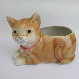 May include: A ceramic planter shaped like a resting orange tabby cat. The cat has a glossy finish, white paws, and a pink collar with a small bow. The open container section is located on the cat's back, featuring a smooth, light-colored interior.