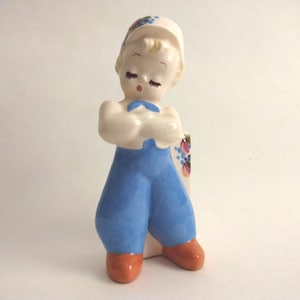 May include: A ceramic figurine of a child wearing a white cap, a white shirt with a blue necktie, and blue overalls. The child has closed eyes and orange shoes. A small floral pattern with pink and blue flowers is visible on the side of the cap.
