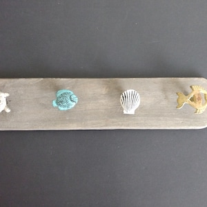 May include: A wooden wall hook with four decorative sea-themed knobs. The knobs include a white turtle, a turquoise fish, a white seashell, and a gold-colored fish. The wooden base is gray and has rounded edges.