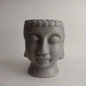 May include: A gray ceramic Buddha head planter with closed eyes and a serene expression. The planter has a detailed hairstyle and a wide opening for plants. The Buddha head is a decorative item for home decor.