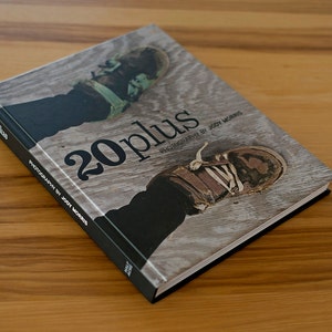 May include: A hardcover book titled "20 plus" by Jody Morris. The cover features a close-up of a pair of worn boots on a wooden background. The book is on a wooden surface.