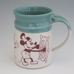 May include: A ceramic mug with a teal glaze and a white design featuring Mickey Mouse and a ship's wheel. Mickey is wearing a captain's hat and holding the wheel.